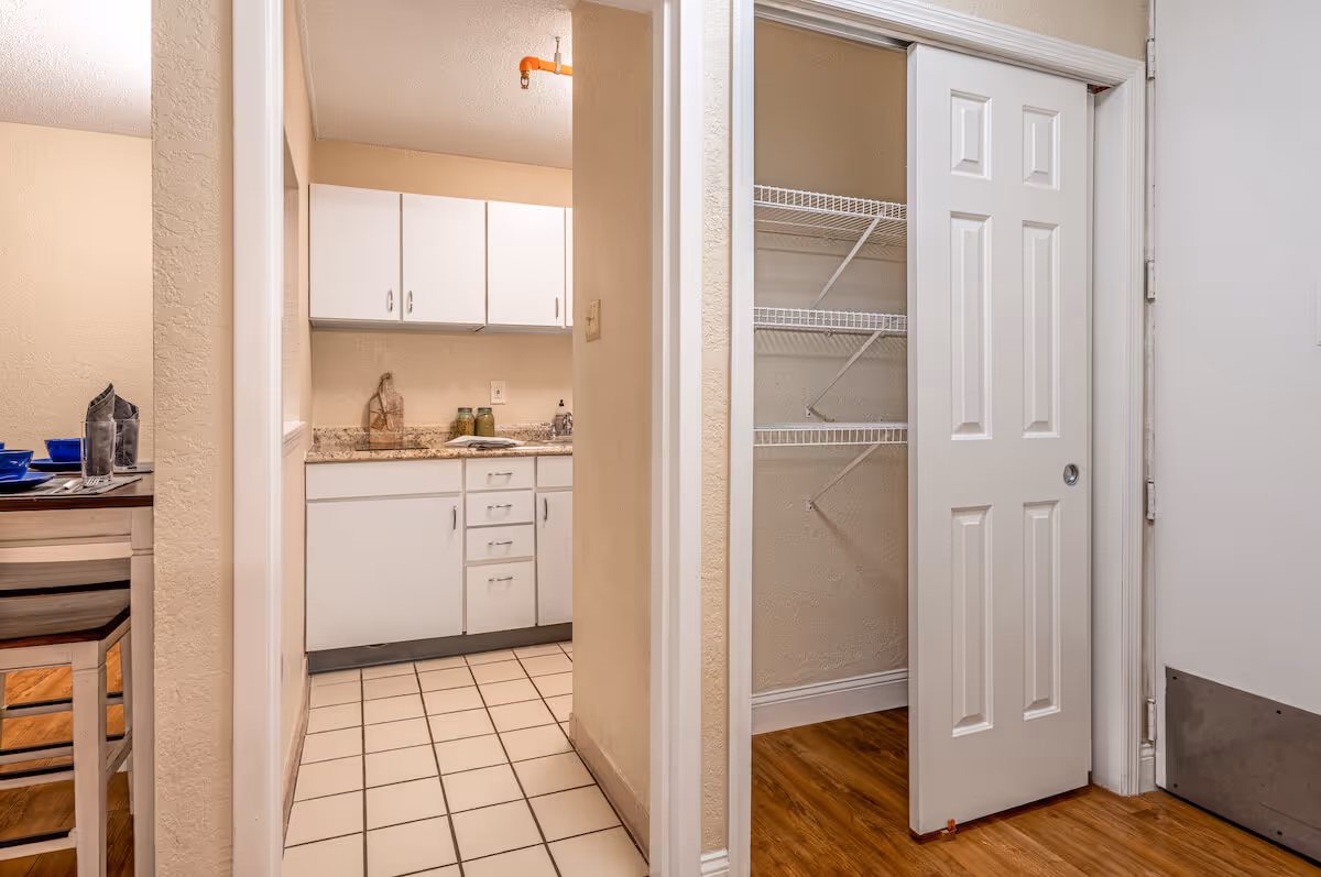 View of a small kitchen with white cabinets and a granite countertop, adjacent to a dining area with a table set with blue dishes. To the right, there is an open closet with white wire shelving and a sliding white door. The floor transitions from white tile in the kitchen to wood flooring in the closet and dining area.