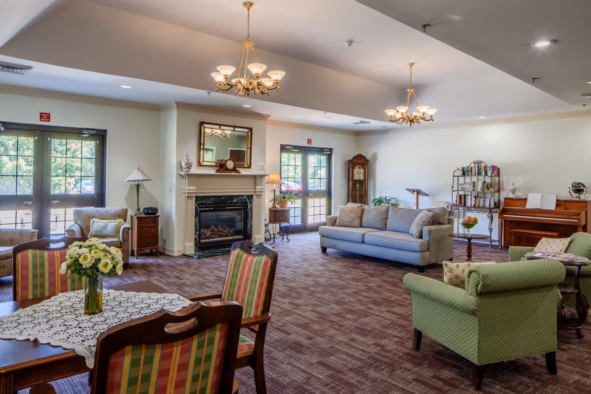 A spacious living room area in a senior living facility with a mix of seating options including a blue sofa, green armchairs, and striped dining chairs around a table with a lace tablecloth and a vase of flowers. The room features a fireplace with a mirror above it, two chandeliers, a grandfather clock, a bookshelf, and a piano. Large windows and glass doors allow natural light to fill the room.