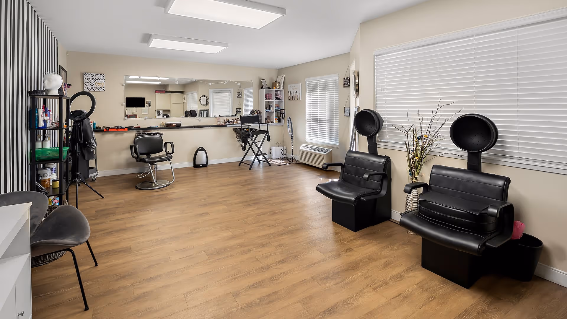 Interior view of a salon area in a senior living facility with wooden flooring, two black hair drying chairs on the right, a black styling chair in front of a large mirror on the left, shelves with hair products and equipment, and large windows with white blinds.