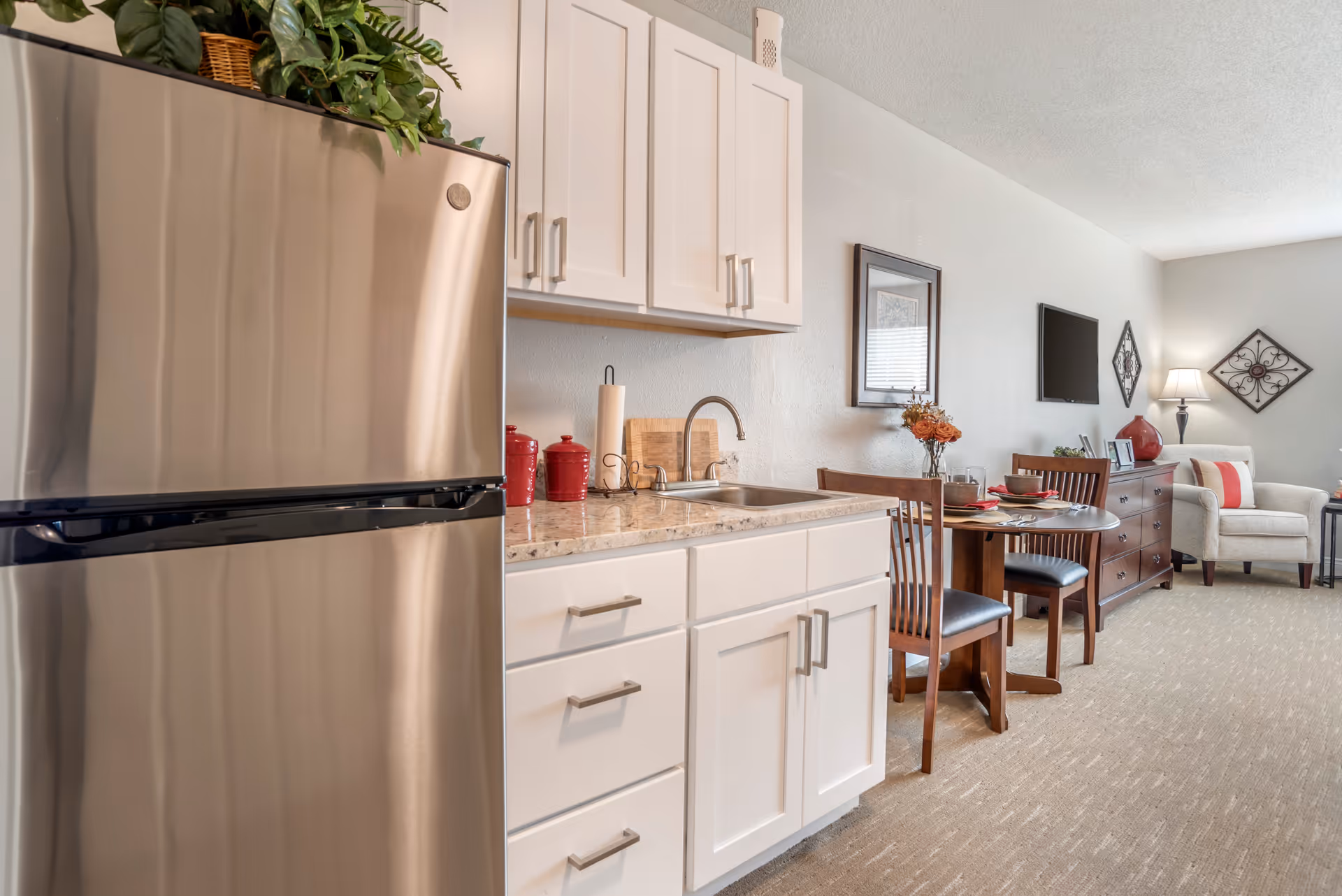 Interior view of a senior living facility apartment featuring a stainless steel refrigerator, white cabinets, a countertop with a sink, and a small dining table set with two chairs. The living area includes a dresser, a wall-mounted TV, an armchair, and decorative wall art and lamp.