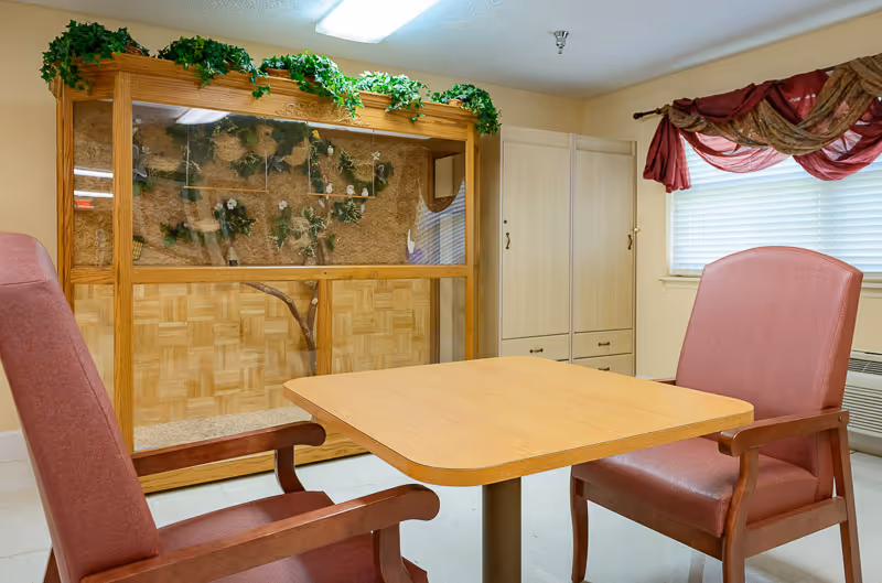 A small room with a wooden table and two upholstered chairs facing each other. Behind the table is a large wooden enclosure with glass front, decorated with green plants and flowers inside. To the right, there is a window with blinds and red curtains, and a beige cabinet against the wall.