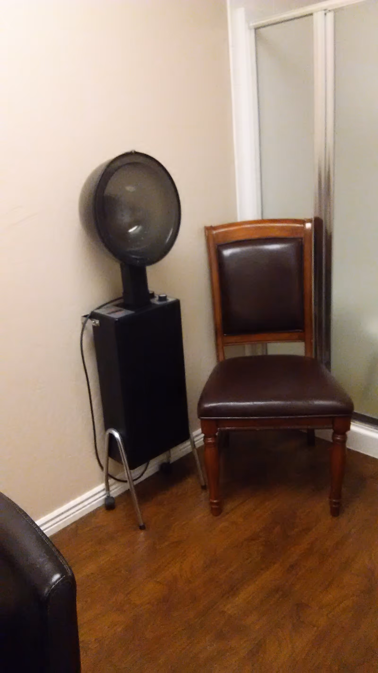 A corner of a room with a brown leather cushioned wooden chair next to a black vintage hair dryer machine on a stand, with a glass shower door in the background and wood flooring.