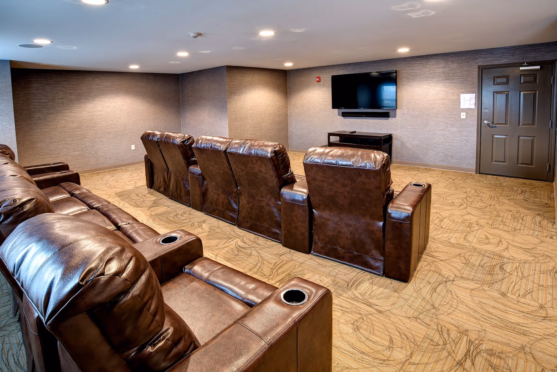 A cozy media room with two rows of brown leather recliner chairs facing a wall-mounted flat screen TV. The room has beige patterned carpet, neutral-colored walls, recessed ceiling lights, and a dark brown door on the right side.
