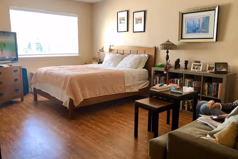 A cozy bedroom with a wooden bed frame and a peach-colored blanket. There is a wooden dresser with a TV on it near a window with blinds. A bookshelf with books, framed photos, and decorative items is against the wall. A small table with two stools is in front of a green sofa where a person is sitting, holding a book or magazine. The room has wooden flooring and light-colored walls with framed artwork.