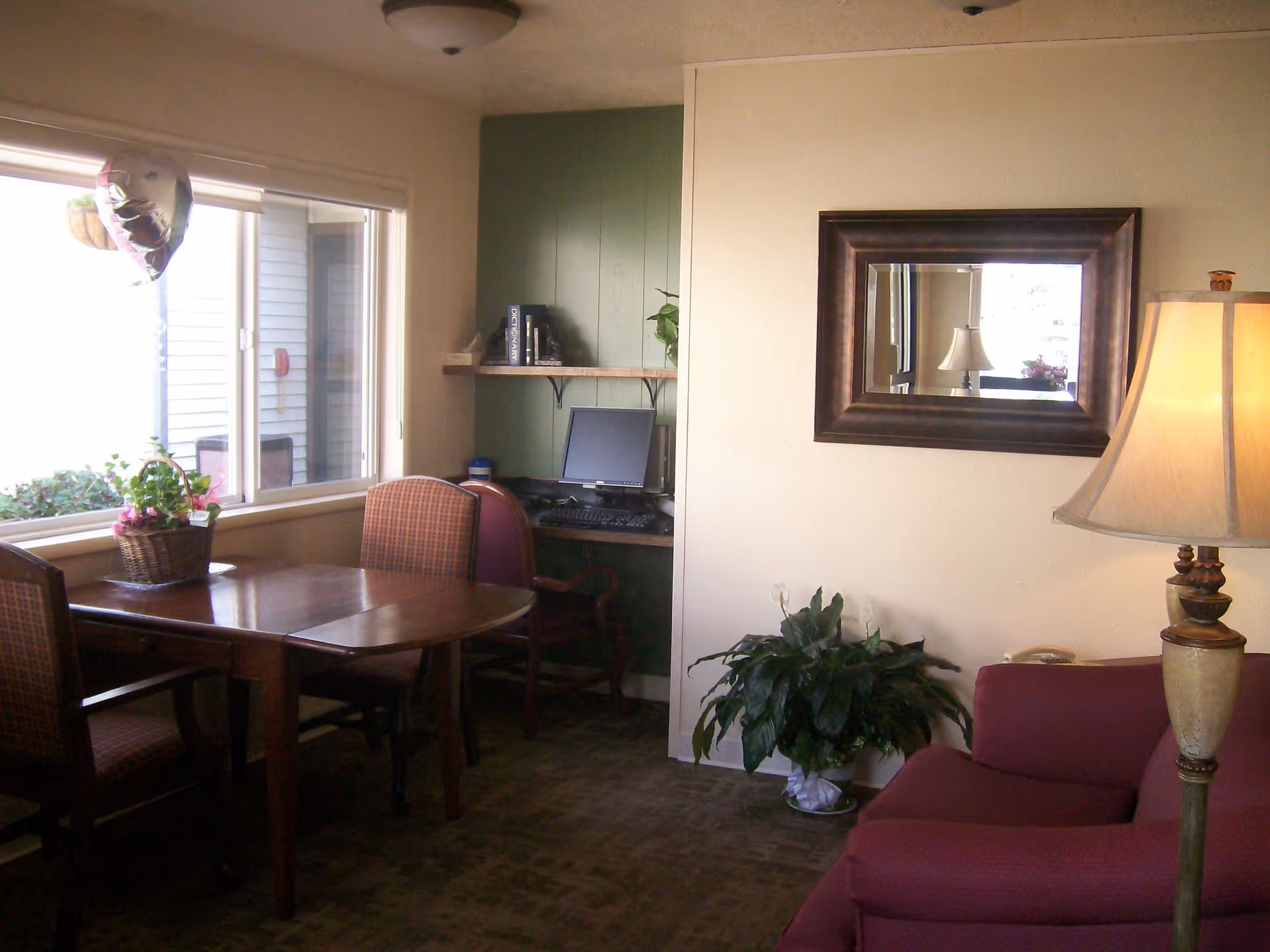 Small communal living room with a table and chairs by a window, a computer desk, potted plants, a mirror, and a lamp.