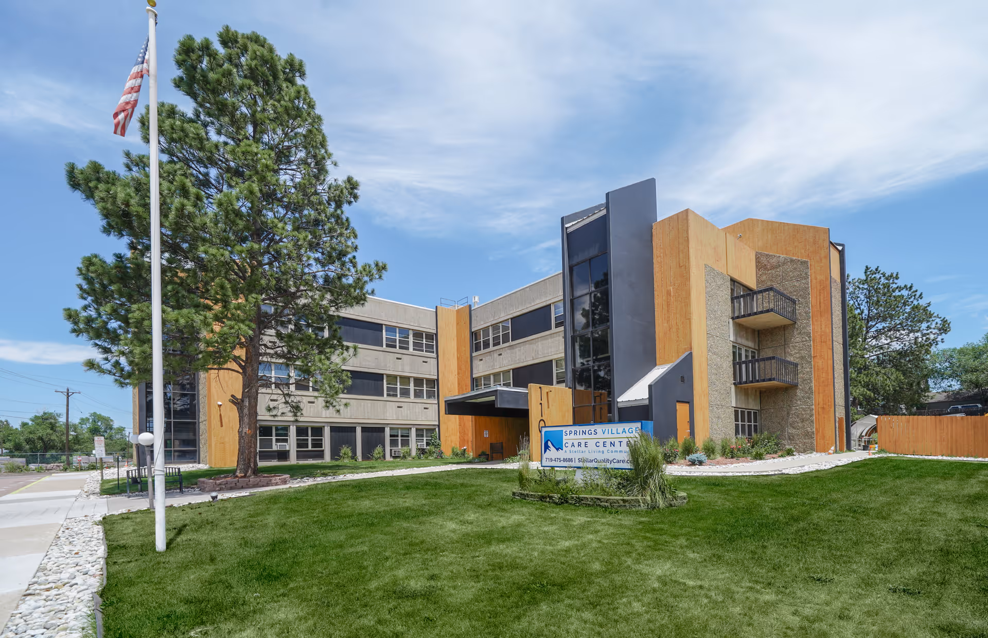 Exterior view of Springs Village Care Center, a three-story building with a modern design featuring large windows and wooden panel accents. There is a green lawn in front with a sign displaying the facility's name and contact information. An American flag is flying on a flagpole to the left, and there are trees and a clear blue sky in the background.
