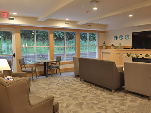 A cozy living room area with large windows showing greenery outside. The room features a patterned carpet, several armchairs, a sofa, a small table with two chairs near the windows, and a TV mounted above a fireplace with decorative vases on the mantel.