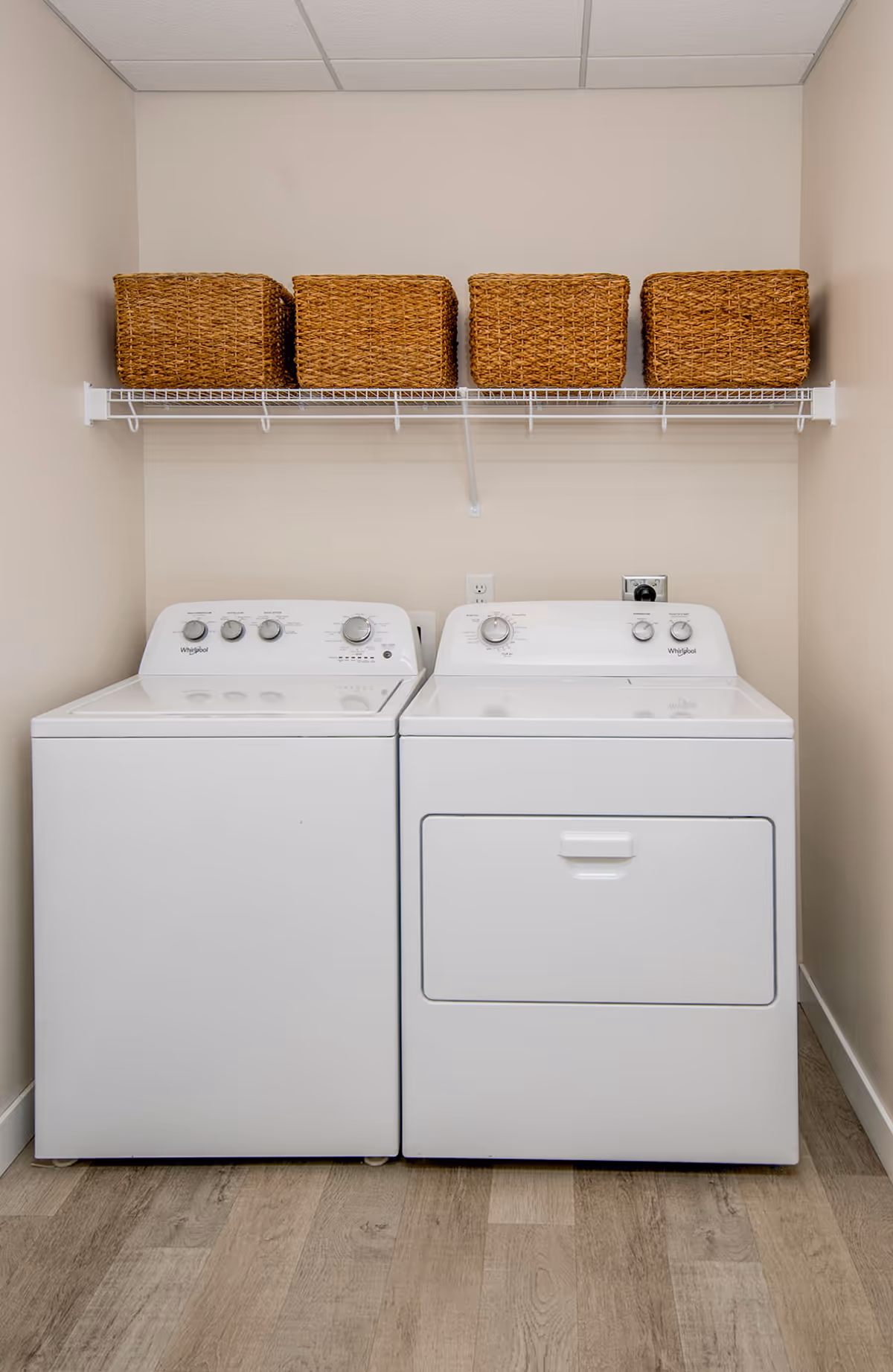 A laundry room with a white washing machine and dryer side by side against a beige wall. Above them is a white wire shelf holding four woven storage baskets. The floor is wood-patterned.