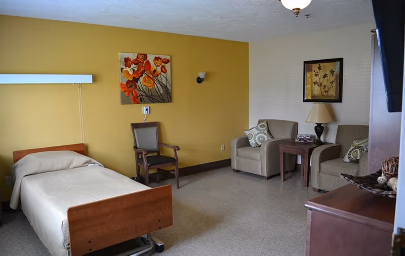 A senior living facility room with a single bed on the left side, a wooden chair next to the bed, and two cushioned armchairs with patterned pillows on the right side. A small wooden side table with a lamp and decorative items is placed between the armchairs. The walls are painted yellow and beige, adorned with floral paintings. The floor is covered with light-colored linoleum.