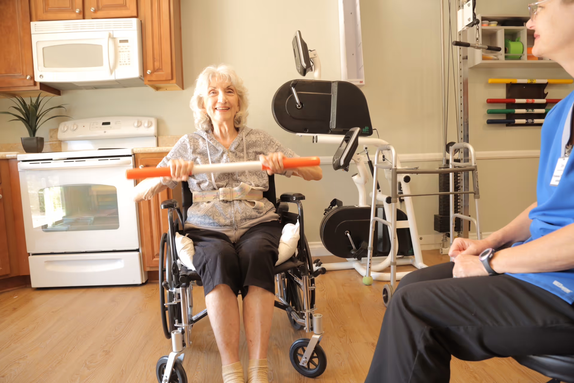An elderly woman in a wheelchair is exercising with a resistance bar in a room that appears to be a kitchen area with exercise equipment in the background. A person in a blue shirt is seated nearby, watching her.