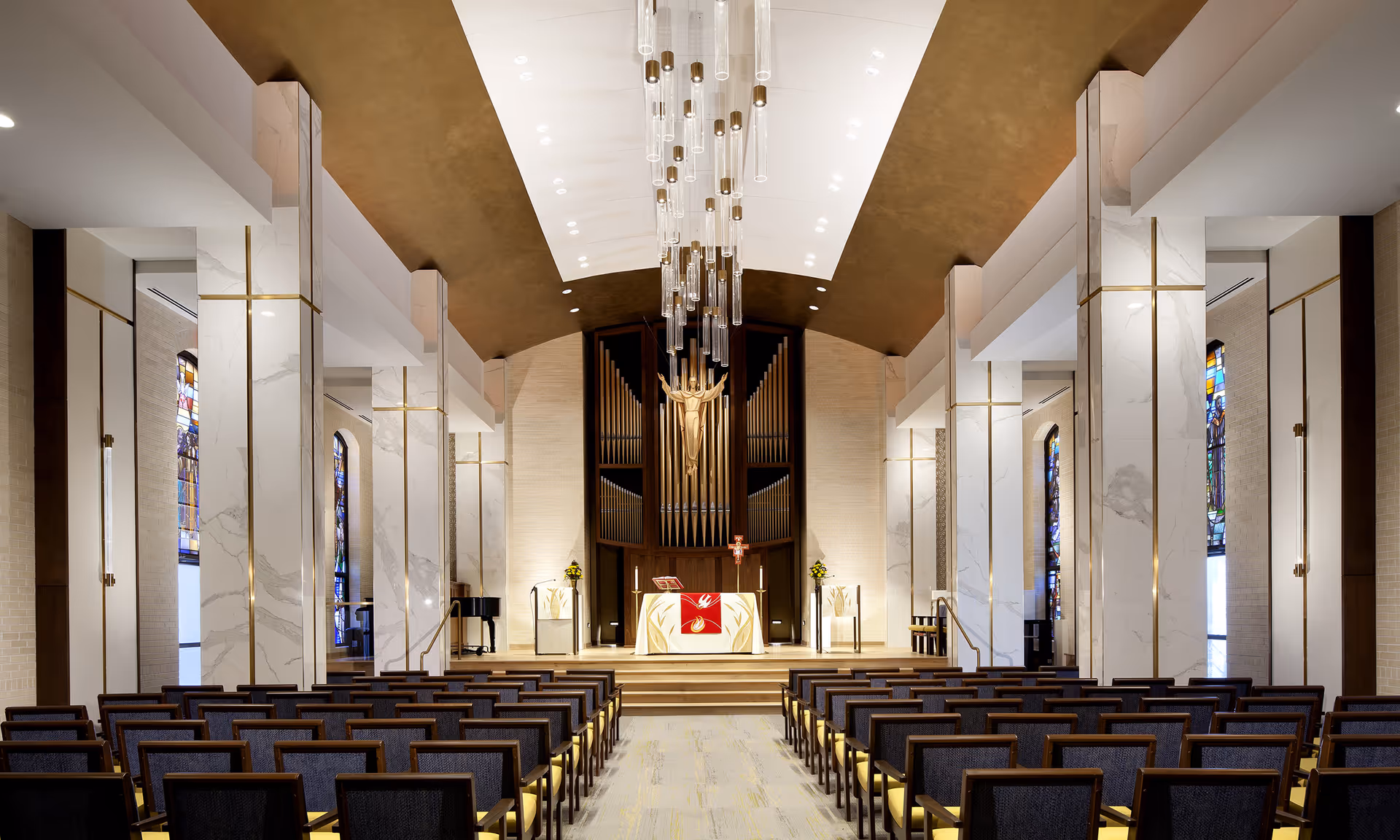 Interior view of a chapel with rows of wooden chairs facing an altar. The altar is decorated with a red and white cloth and flanked by two stands with candles and flowers. Behind the altar is a large pipe organ and a golden statue of Jesus with outstretched arms. The ceiling features a modern chandelier with multiple hanging glass tubes. Stained glass windows line the side walls.