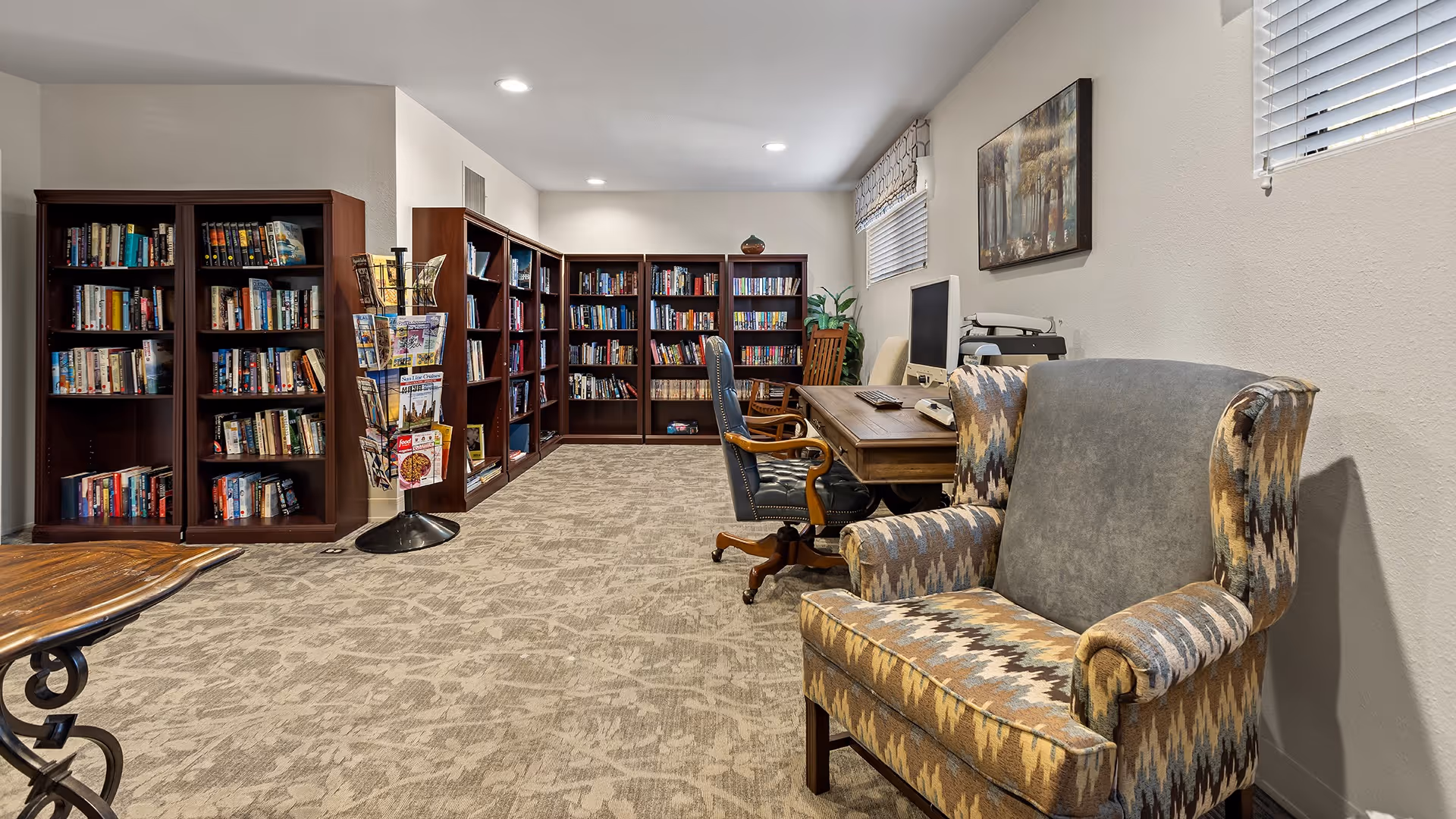 A cozy library or reading room with multiple dark wood bookshelves filled with books along the walls. There is a patterned armchair in the foreground, a wooden desk with two office chairs, a computer, and a printer. The room has carpeted flooring, neutral-colored walls, and two windows with blinds and valances.
