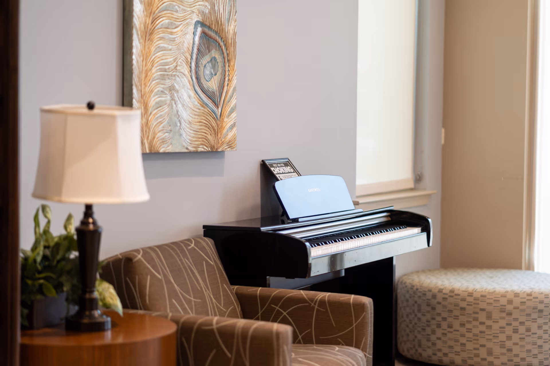 Cozy lounge area with an upright piano, patterned armchair, side table with lamp and a piece of wall art.