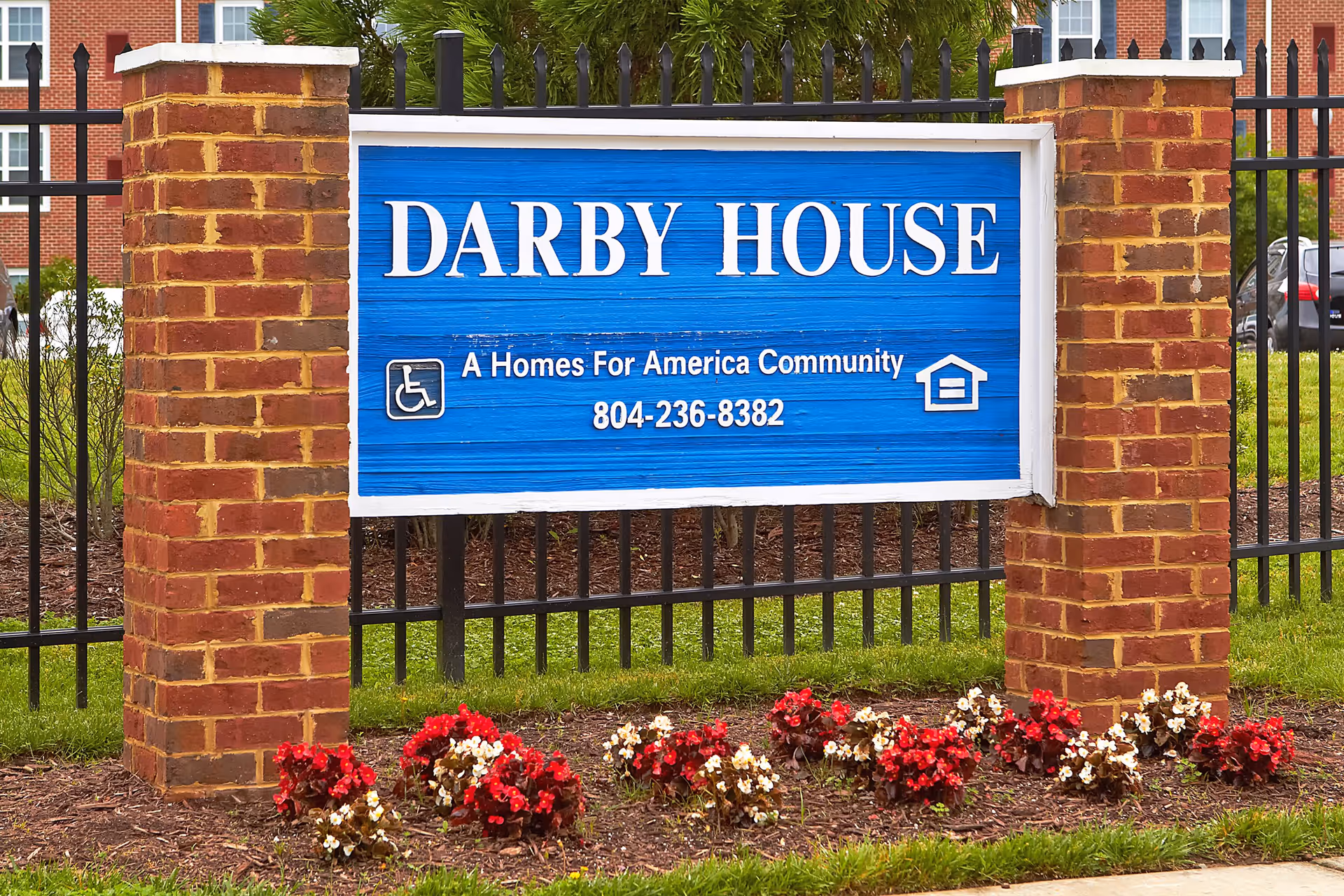 A blue sign with white text reading 'DARBY HOUSE A Homes For America Community 804-236-8382' mounted between two brick pillars with a black metal fence behind it and red and white flowers planted in front.