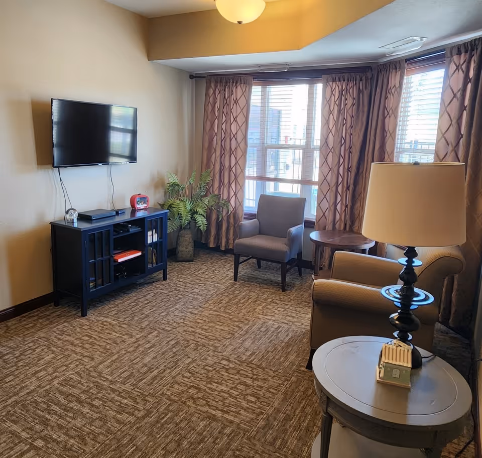 Cozy living room with armchairs, a wall-mounted TV over a media cabinet, side tables, a lamp, and bay windows with curtains.
