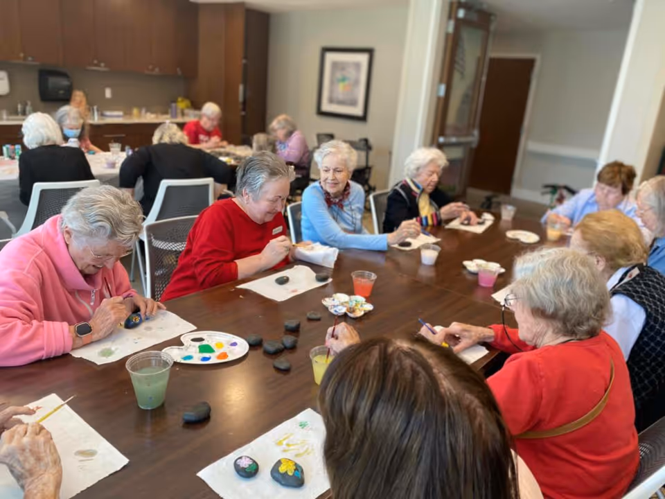 A group of elderly women seated around a large table engaged in painting small rocks with colorful designs. The room has a warm, communal atmosphere with cabinets and a countertop in the background.