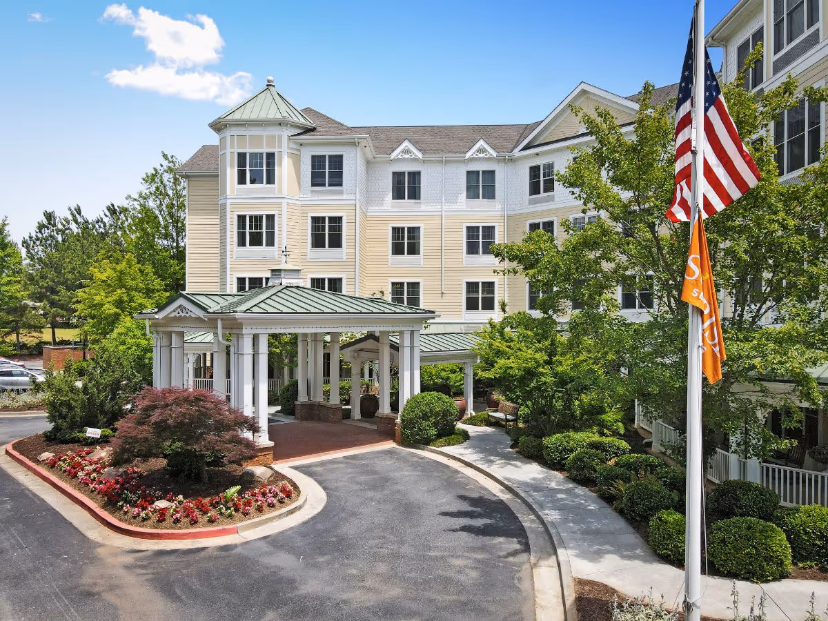 Exterior view of Sunrise of Johns Creek senior living facility showing a multi-story building with beige and white siding, a covered entrance with white columns and a green roof, landscaped bushes and flowers, and an American flag along with an orange Sunrise flag on a flagpole.