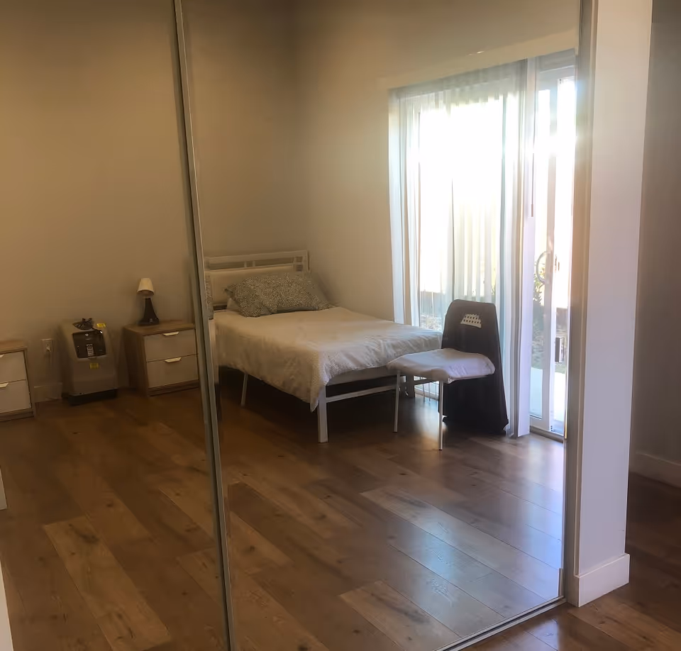 A simple bedroom with a single bed covered with a white blanket and a patterned pillow. Next to the bed is a small chair with a cushion and a dark cloth draped over it. There are two small wooden nightstands, one with a lamp and the other with a small device. The room has wooden flooring and a large sliding glass door with vertical blinds letting in natural light. A large mirror reflects the room.