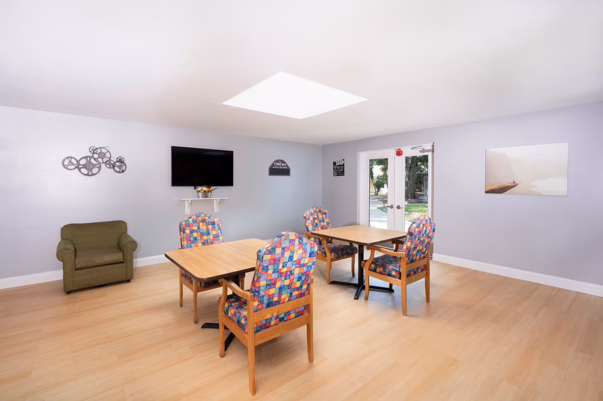 A bright room with light wooden flooring and light gray walls featuring two wooden tables with colorful patterned chairs around them. There is a green armchair in the corner, a flat-screen TV mounted on the wall, and double glass doors leading outside. The room is decorated with a metal wall art piece, a small shelf under the TV, a cinema-themed sign, and a landscape painting on the walls.