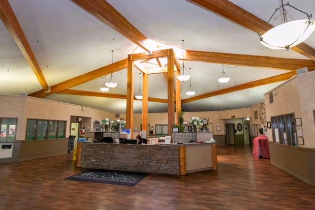 Spacious reception area with a central desk made of stone and wood, featuring a person seated behind it. The ceiling has exposed wooden beams and multiple hanging light fixtures. The floor is wooden, and there are several doors and windows along the walls. The area is well-lit and decorated with plants and flowers.