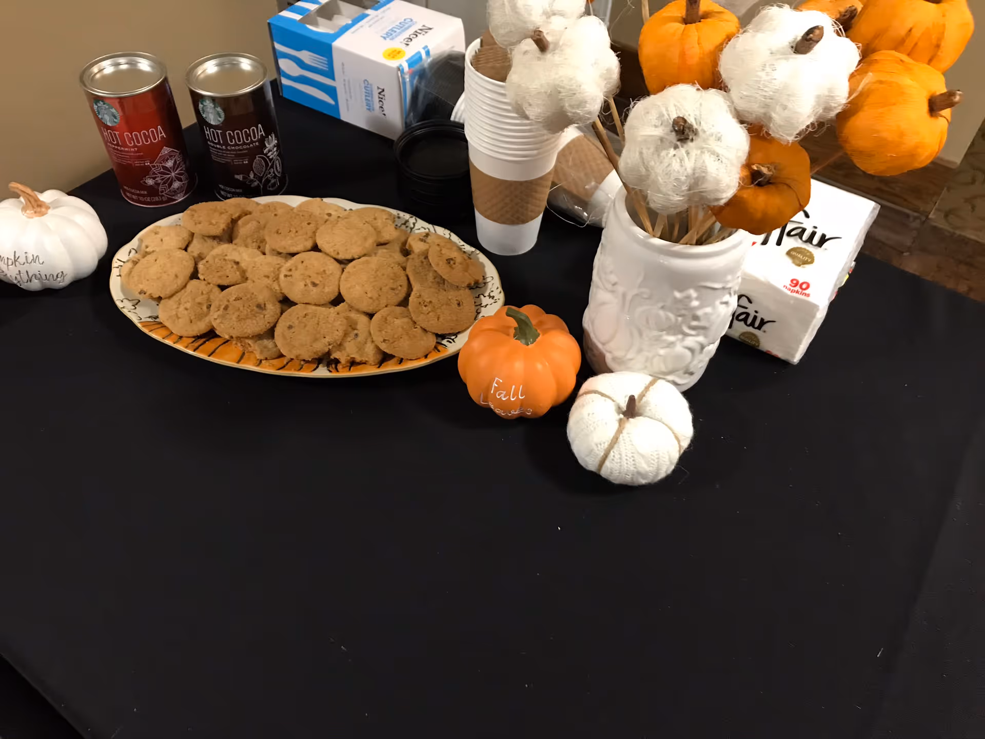 Table with a plate of cookies, hot cocoa cans, paper cups and fall pumpkin decorations on a black tablecloth.