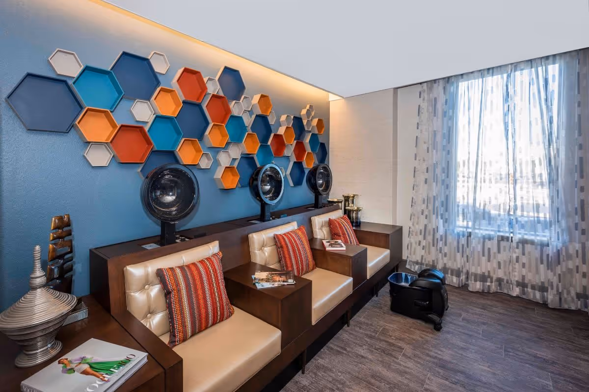 A bright salon-style lounge with three hooded hair dryers, cushioned chairs with colorful pillows, hexagonal wall decor, and a sheer-curtained window.