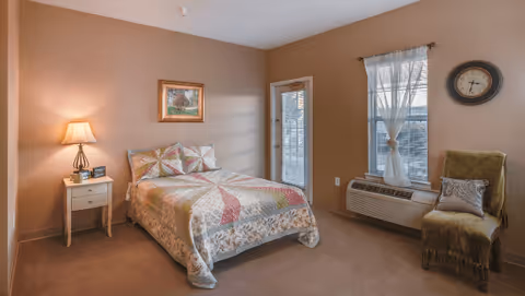 A cozy bedroom with a double bed covered in a patterned quilt, a nightstand with a lamp and clock, a framed painting on the wall, a window with sheer curtains, a wall clock, an air conditioning unit below the window, and a green upholstered chair with a decorative pillow.