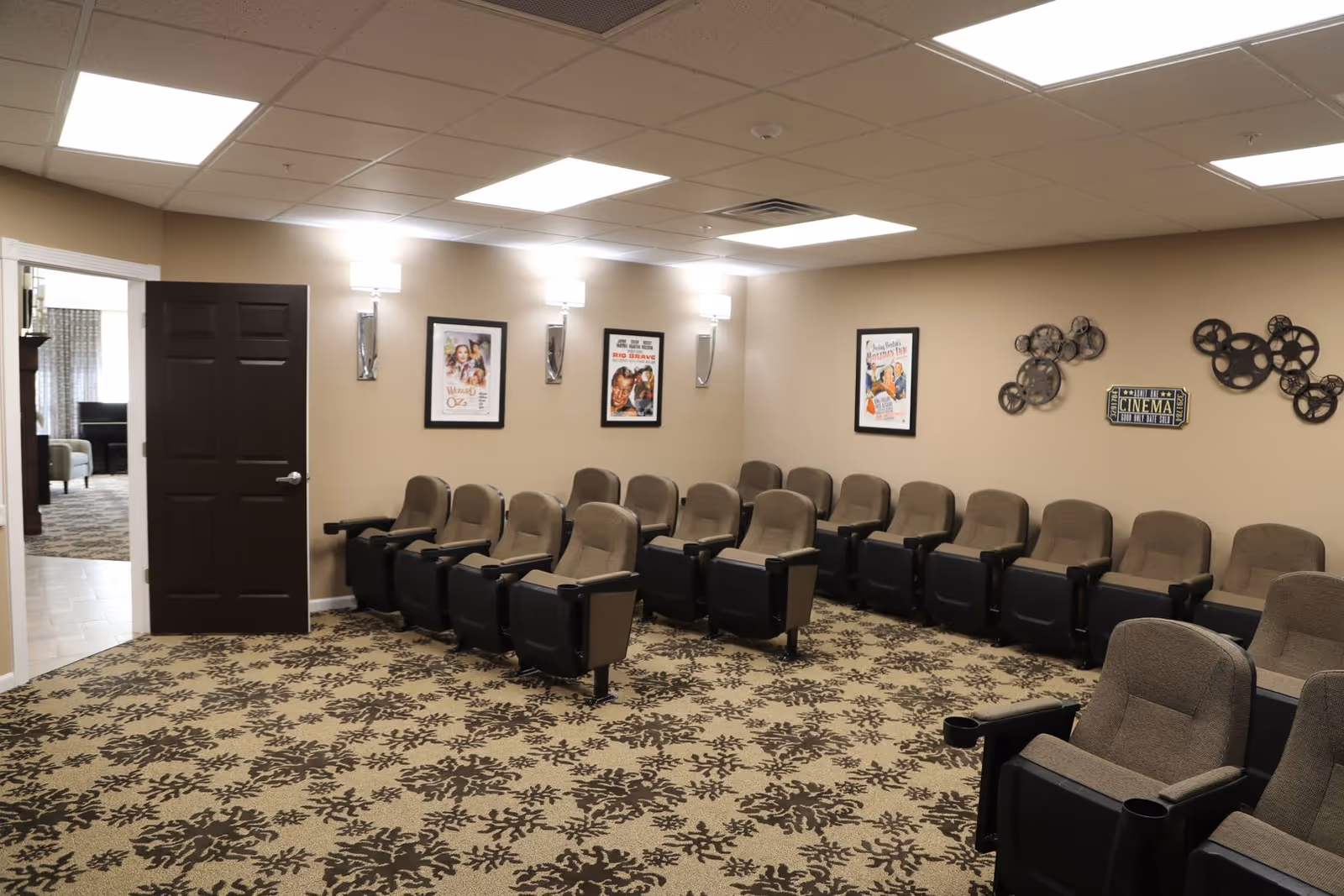 A small movie theater room with two rows of cushioned theater seats arranged in front of a beige wall. The wall is decorated with framed vintage movie posters and metal film reel art. The room has patterned carpet flooring and bright ceiling lights. An open door leads to another room with a piano and seating area.
