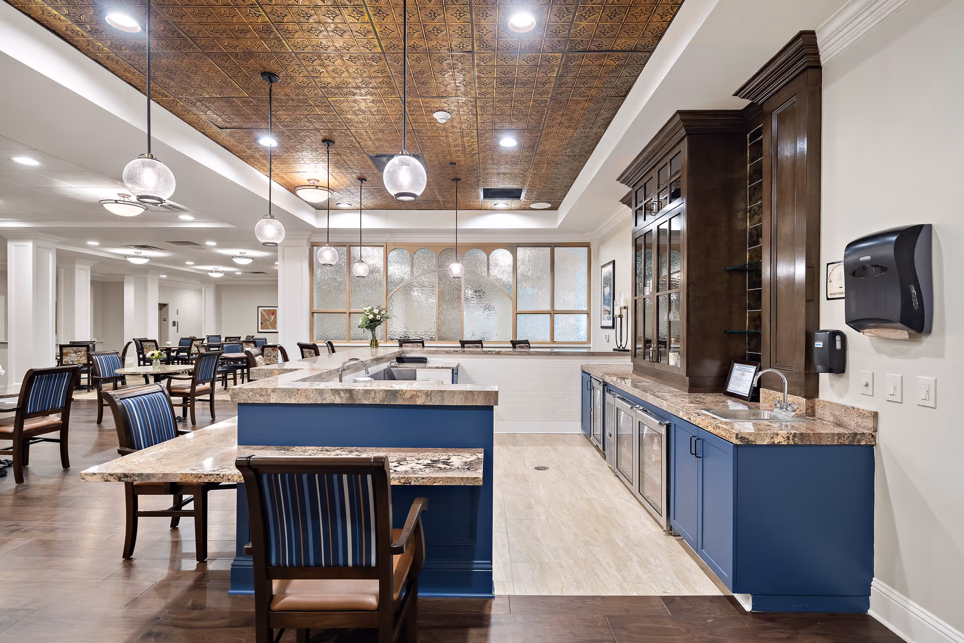 A spacious dining area in Worthington Manor featuring multiple tables and chairs with blue-striped upholstery. The room has a decorative ceiling with pendant lights hanging above a central counter area with a marble countertop and blue cabinetry. There is a wooden cabinet with glass doors and a sink on the right side, and frosted glass panels in the background.