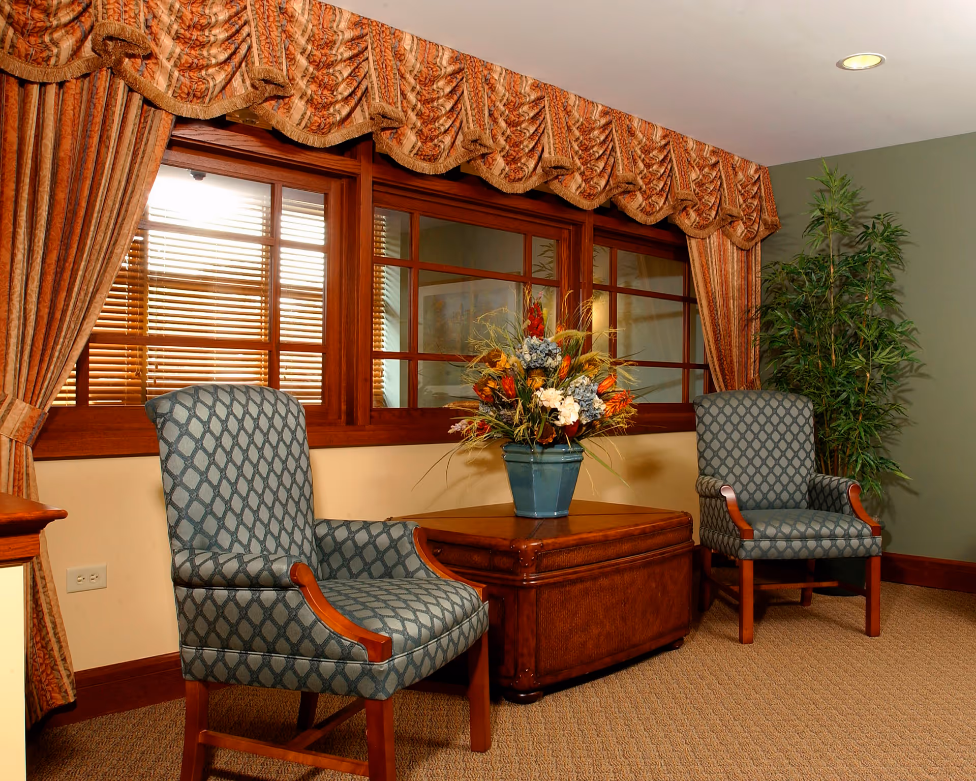 A cozy sitting area with two patterned armchairs facing each other and a wooden coffee table between them holding a floral arrangement. Behind the chairs is a large window with wooden blinds and ornate draped curtains. A tall green plant is placed in the corner of the room.