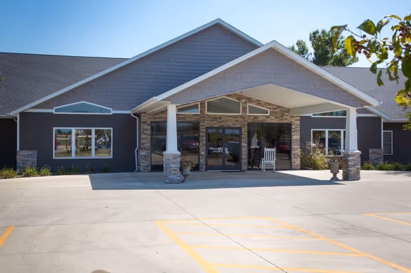 Front entrance of High Point Residence Taylorville with a covered porte-cochere, stone pillars, glass doors and a parking area.