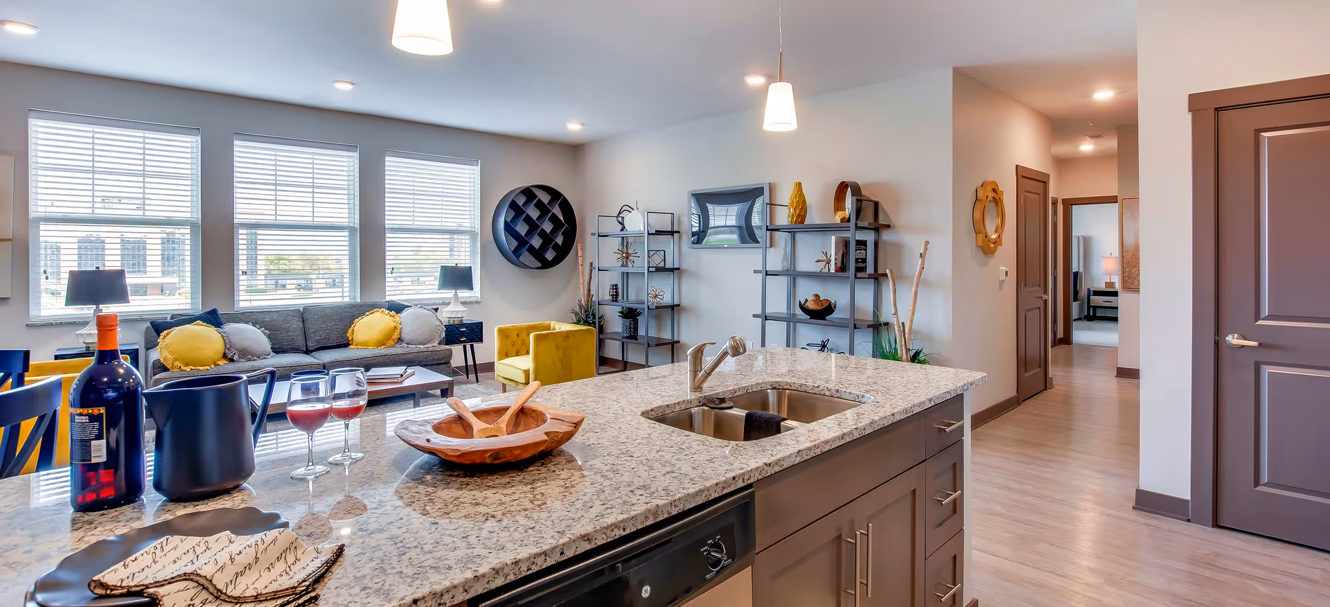 Open-concept apartment living area and kitchen with a granite island and sink, sofa, armchair, shelving, and large windows.