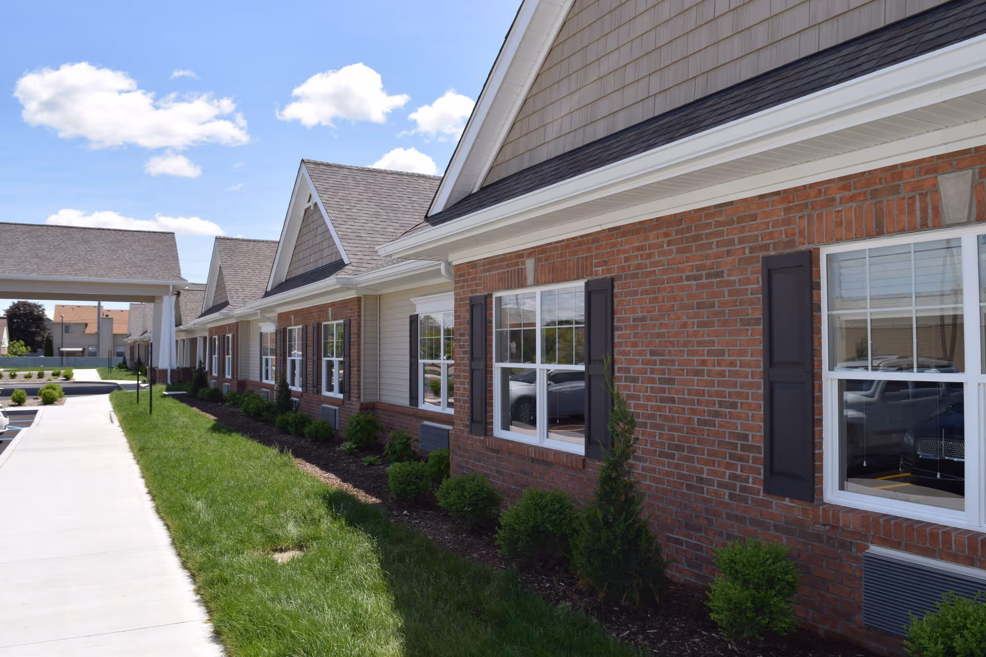 Brick and siding exterior of a one-story assisted living building with windows, shrubs, and a sidewalk leading to a covered entrance.