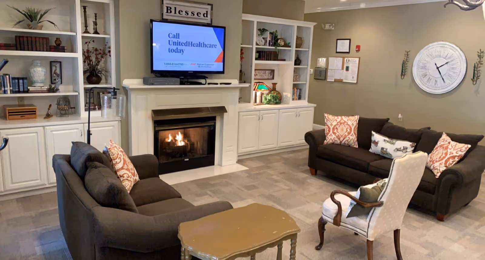 Cozy common living room with sofas arranged around a fireplace and wall-mounted TV, built-in shelves, and a large wall clock.