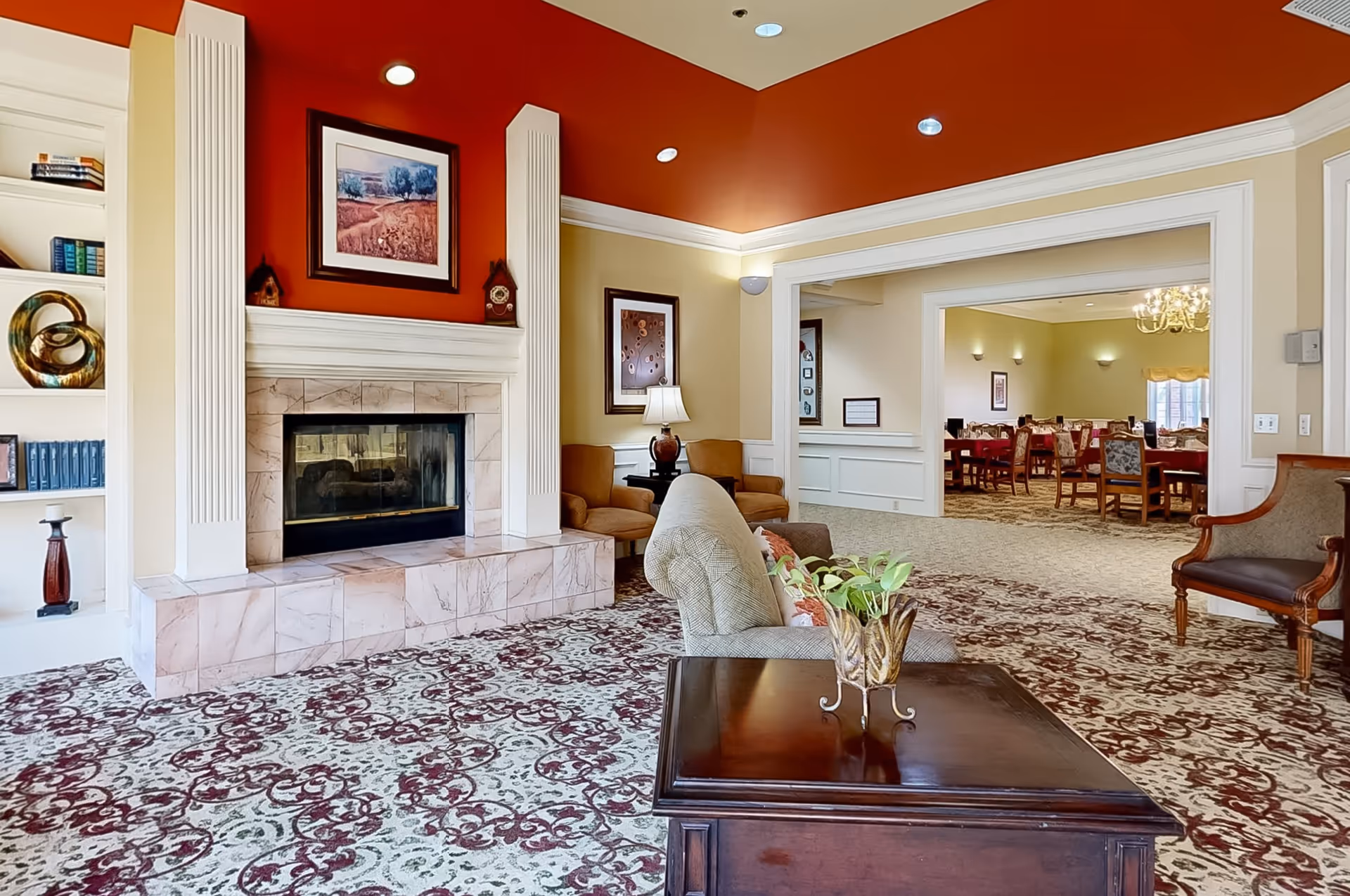 A cozy living room area with a patterned carpet, a marble fireplace with a red accent wall above it, and framed artwork. The room has comfortable seating including a sofa and chairs, a wooden coffee table with a decorative plant, and a view into a dining room with multiple tables and chairs.