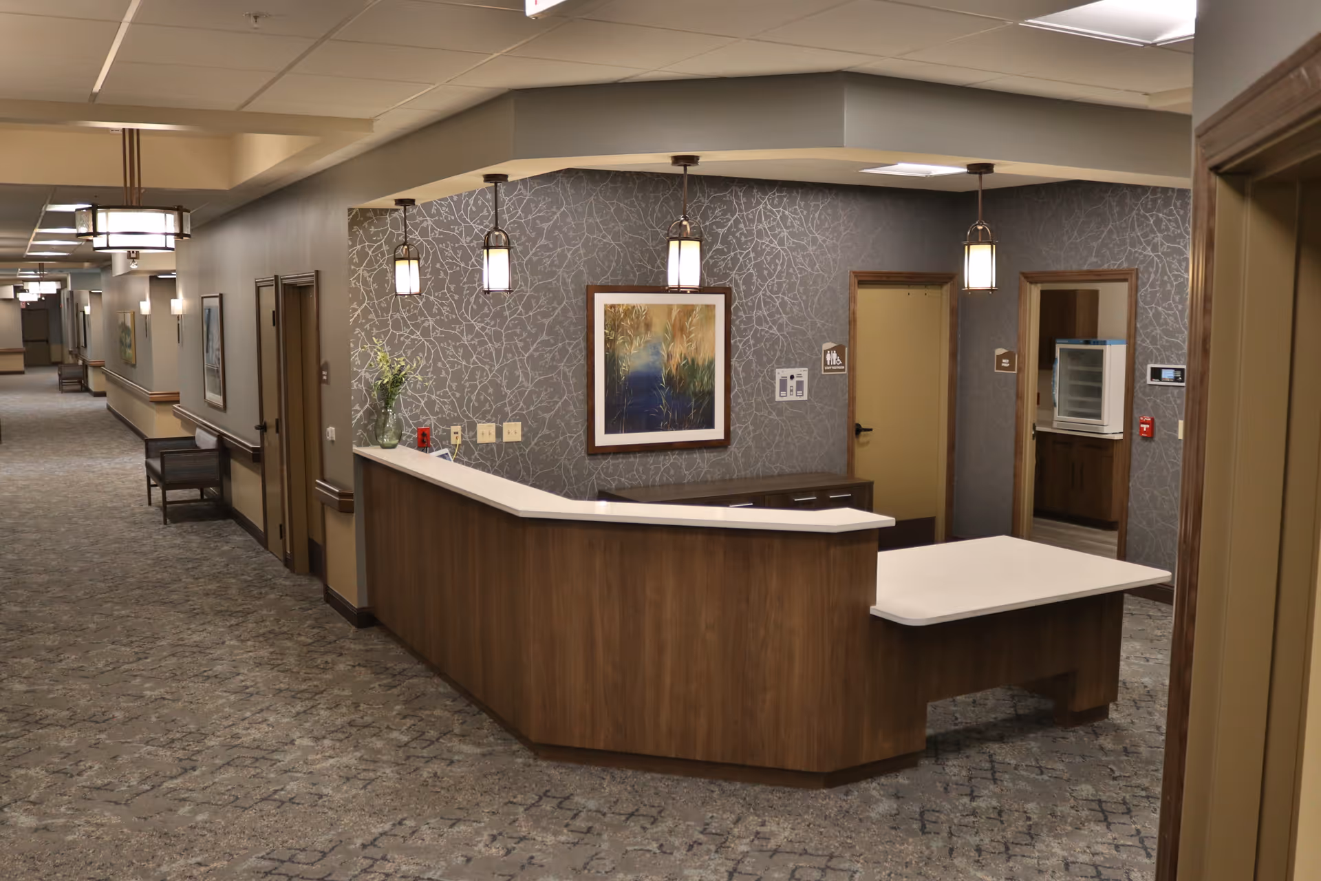 Interior view of a senior living facility hallway with a wooden reception desk featuring a white countertop. The wall behind the desk has a textured gray wallpaper with a framed artwork and three pendant lights hanging above. There are doors and a small room visible behind the desk, and the hallway extends into the distance with chairs and wall-mounted lights.