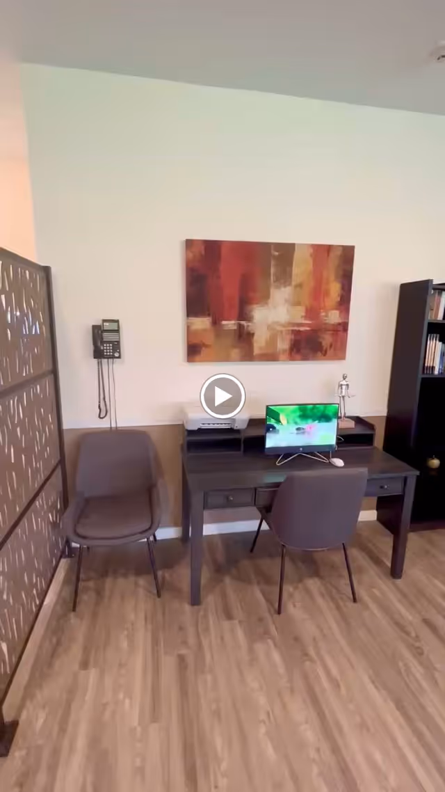 Small interior office area with a desk, two chairs, a computer monitor, and abstract wall art on the wall.