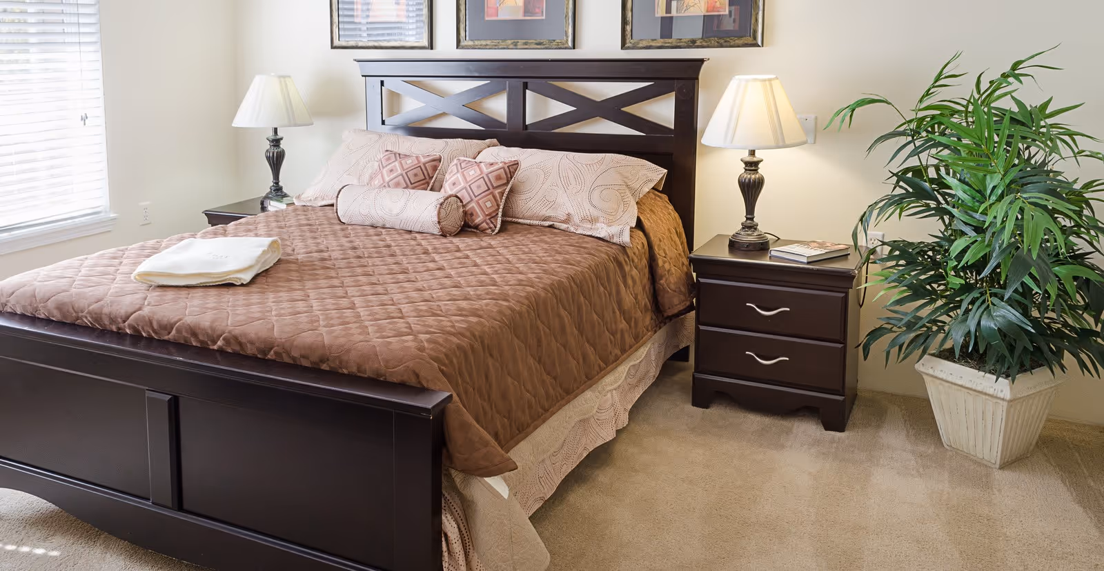Well-lit bedroom with a dark wood bed dressed in a brown quilted bedspread, matching nightstands and lamps, framed art above the headboard, and a potted plant.