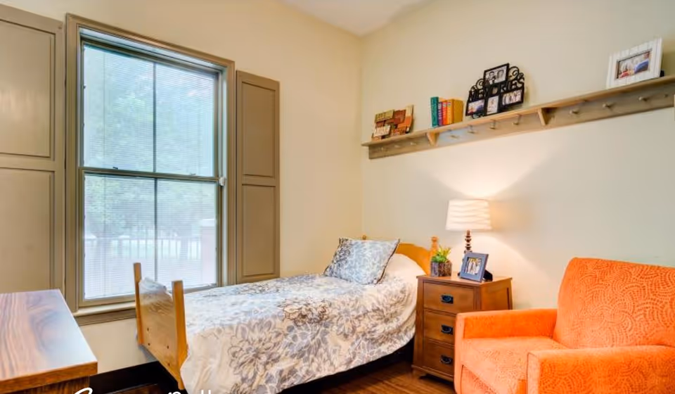 A cozy bedroom with a single wooden bed covered with a floral patterned bedspread, a wooden nightstand with a lamp, framed photos, and a small plant. There is an orange upholstered armchair next to the nightstand. A large window with closed blinds is on the left wall, and a wooden shelf with books and decorative items is mounted on the wall above the bed.