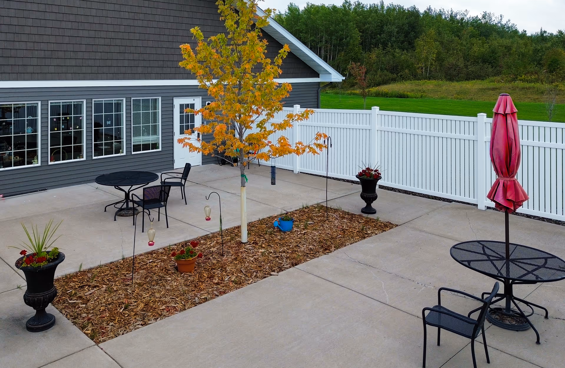 Outdoor patio area with concrete flooring, featuring black metal tables and chairs. One table has a closed red umbrella. There is a small garden bed with a young tree and several potted plants. The patio is adjacent to a gray building with multiple windows and a white door, and is enclosed by a white fence. Green grass and trees are visible beyond the fence.