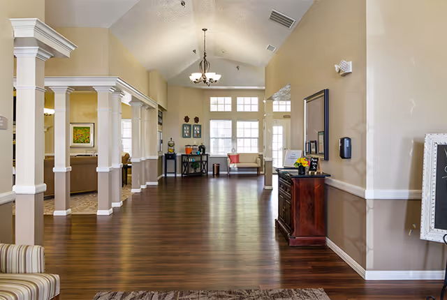 Spacious interior hallway of a senior living facility with wooden flooring, beige walls, white columns, and high ceilings. There are seating areas with chairs and a small table near large windows letting in natural light. A chandelier hangs from the ceiling and decorative items are placed on furniture along the walls.