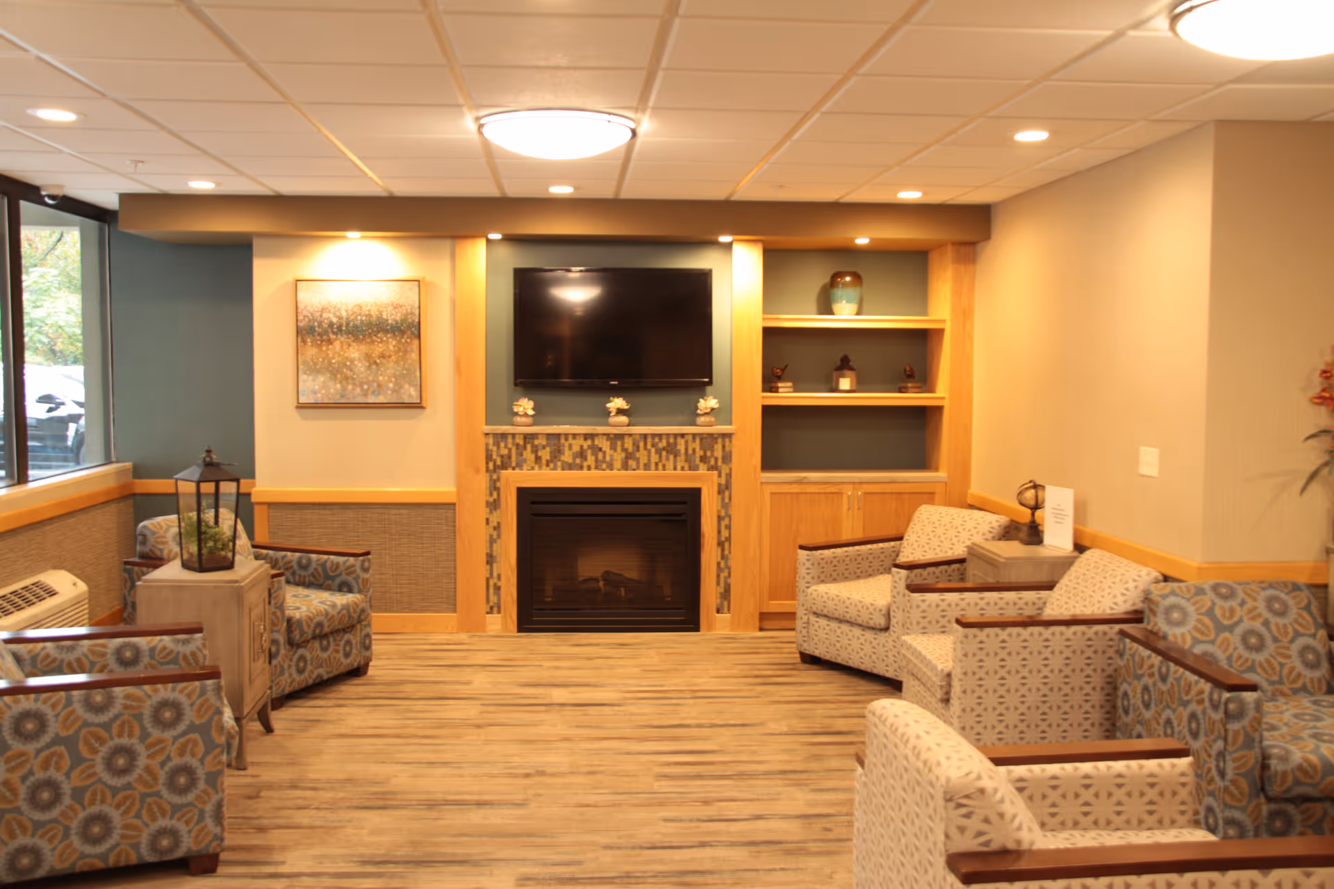 A cozy common area with patterned armchairs arranged around a fireplace with a mounted flat-screen TV above it. The room features built-in wooden shelves with decorative items, a large window letting in natural light, and warm wood and neutral tones throughout.