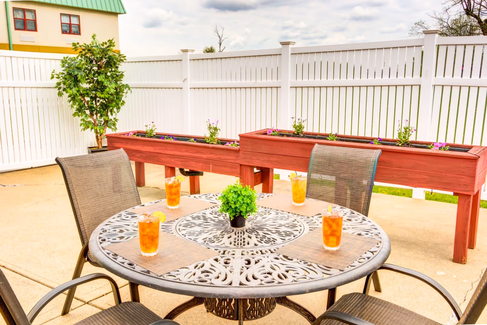 Outdoor patio area with a round metal table and four mesh chairs. On the table are four glasses of iced tea with lemon slices and a small green potted plant in the center. Behind the table are two raised wooden planter boxes with flowers and a small tree in a pot. The area is enclosed by a white vinyl fence and a building with a green roof is partially visible in the background.