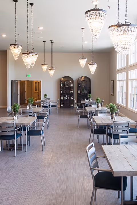 Bright dining room with multiple tables and chairs, hanging crystal chandeliers, large windows, and a wine cabinet at the far wall.