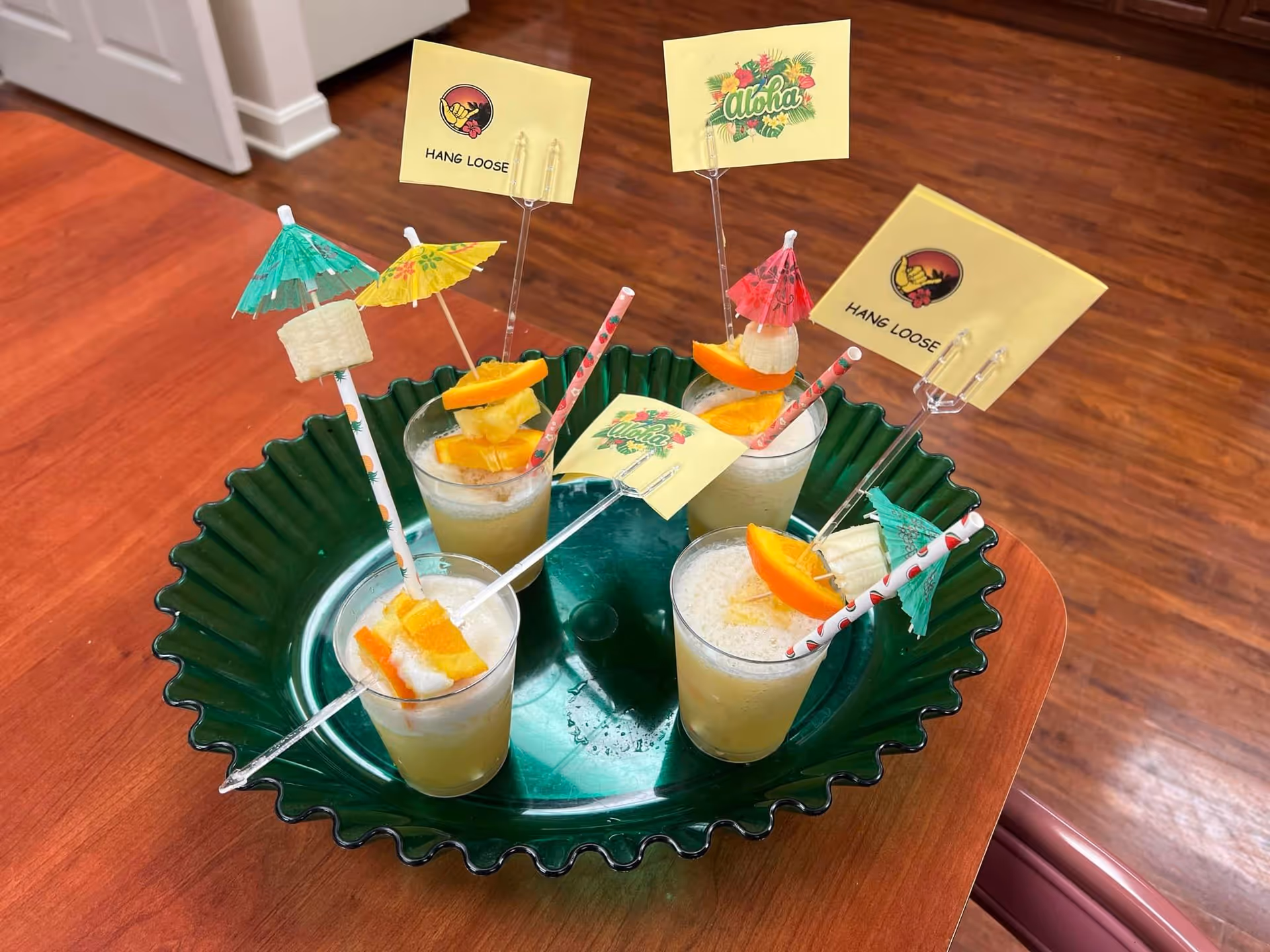 A green tray holding five tropical-themed drinks garnished with orange slices, pineapple chunks, small paper umbrellas, and decorative stirrers with flags that say 'Hang Loose' and 'Aloha'. The tray is placed on a wooden table with a hardwood floor in the background.