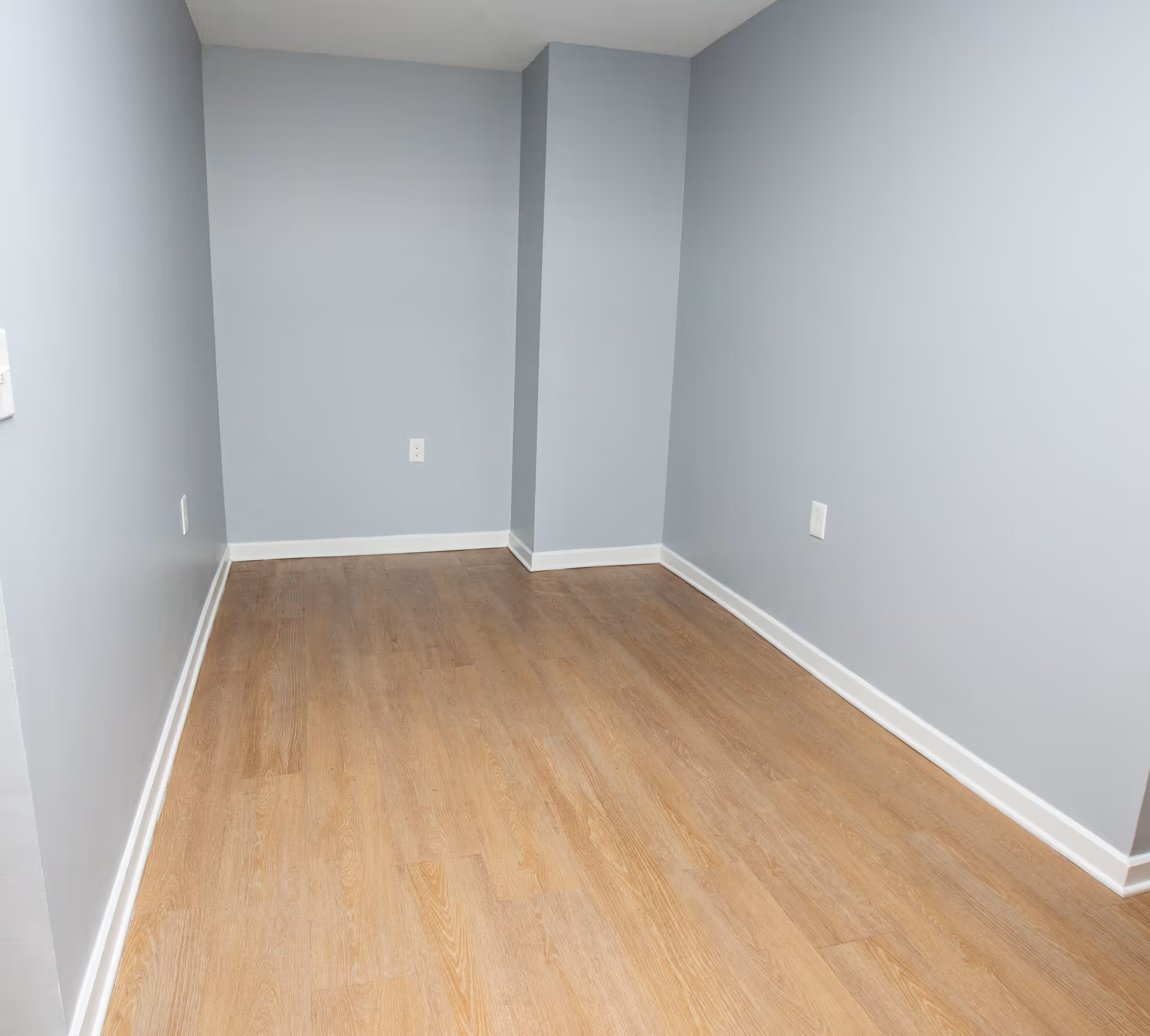 Empty narrow room with light gray walls, light wood flooring, white baseboards, and electrical outlets on the walls.