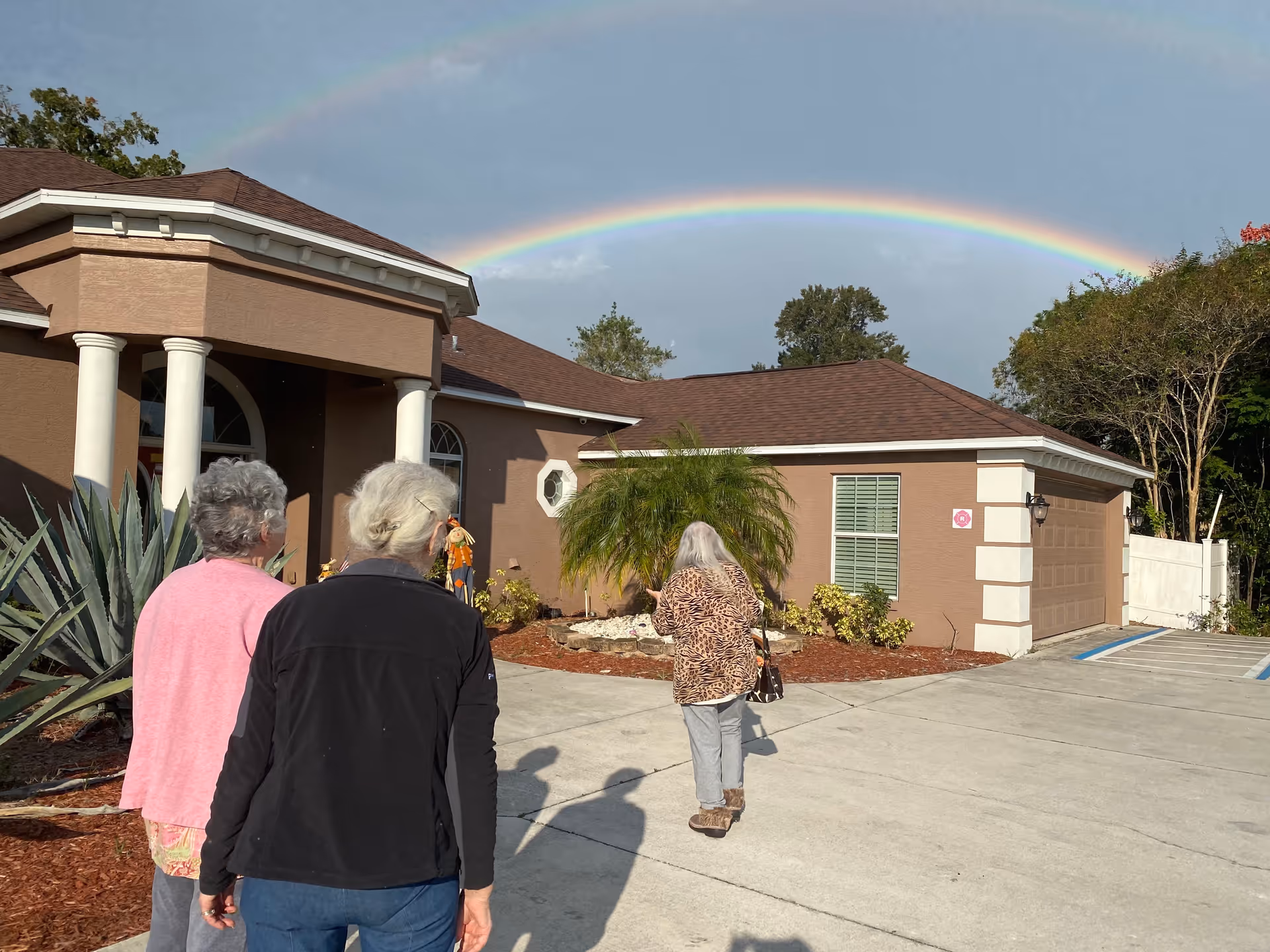 Three elderly women walking towards the entrance of a brown building with white columns under a bright sky with a visible rainbow.