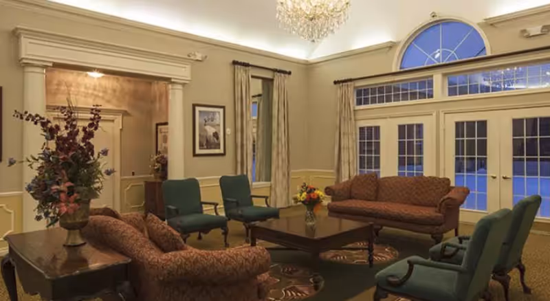 A cozy living room with two patterned sofas and four green armchairs arranged around a dark wooden coffee table with a flower vase. The room features large windows with curtains, a chandelier, and a side table with a floral arrangement.