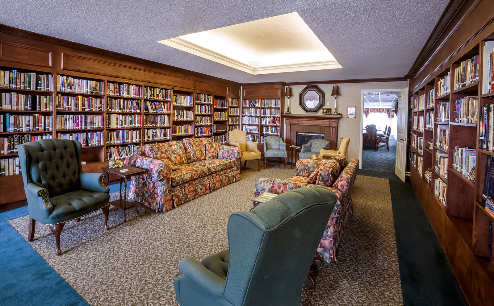 Cozy library-style common room with bookshelves lining the walls, floral sofas, green armchairs, and a fireplace.