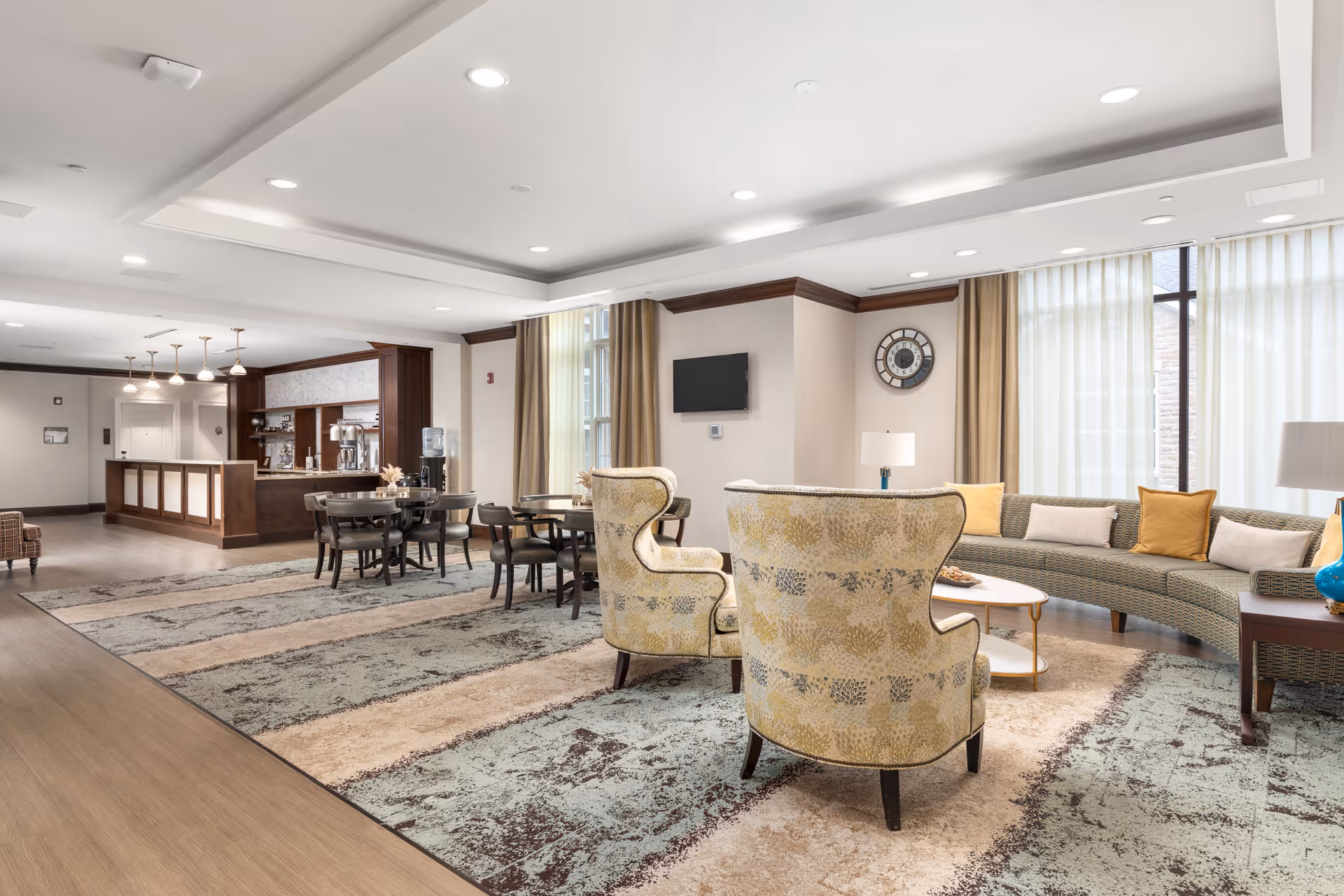 A spacious and well-lit assisted living facility common area with a curved sofa adorned with yellow and white cushions, two patterned armchairs, a coffee table, and several dining tables and chairs. The room features large windows with sheer curtains, a wall clock, a mounted flat-screen TV, and a wooden counter area with pendant lighting in the background.